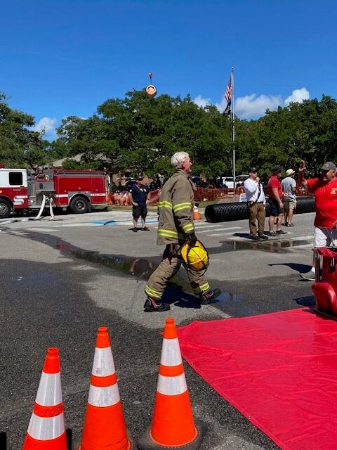 An SJFD firefighter in full turnout gear walks the competition course carrying a yellow helmet