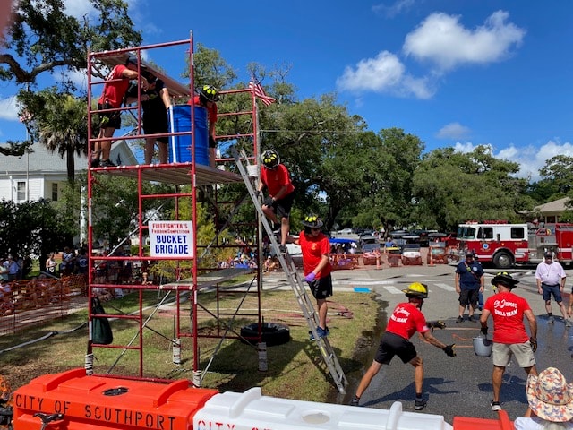 SJFD volunteers scaling the Bucket Brigade scaffold with the competition course and fire trucks visible in the background