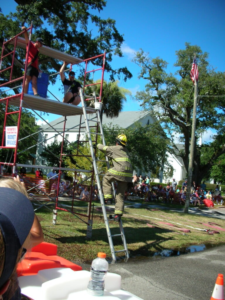 Firefighter in full turnout gear climbing the Bucket Brigade scaffold ladder with a crowd of spectators watching