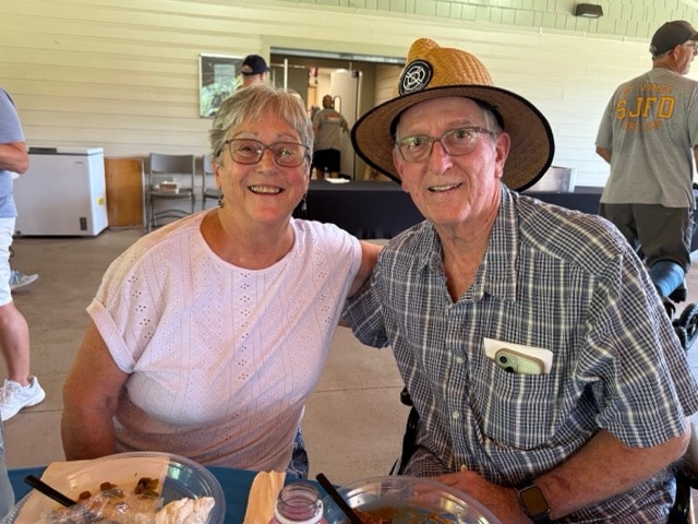 SJFD volunteer couple smile together at the picnic table, the gentleman wearing a straw hat