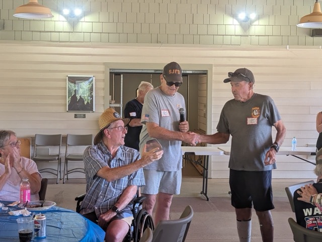 Garren Foy in an SJFD cap with microphone presents a commemorative award to a long-serving volunteer seated in a straw hat at the picnic pavilion