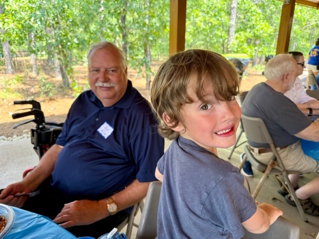 A young boy grins at the camera while seated next to an SJFD volunteer at the picnic