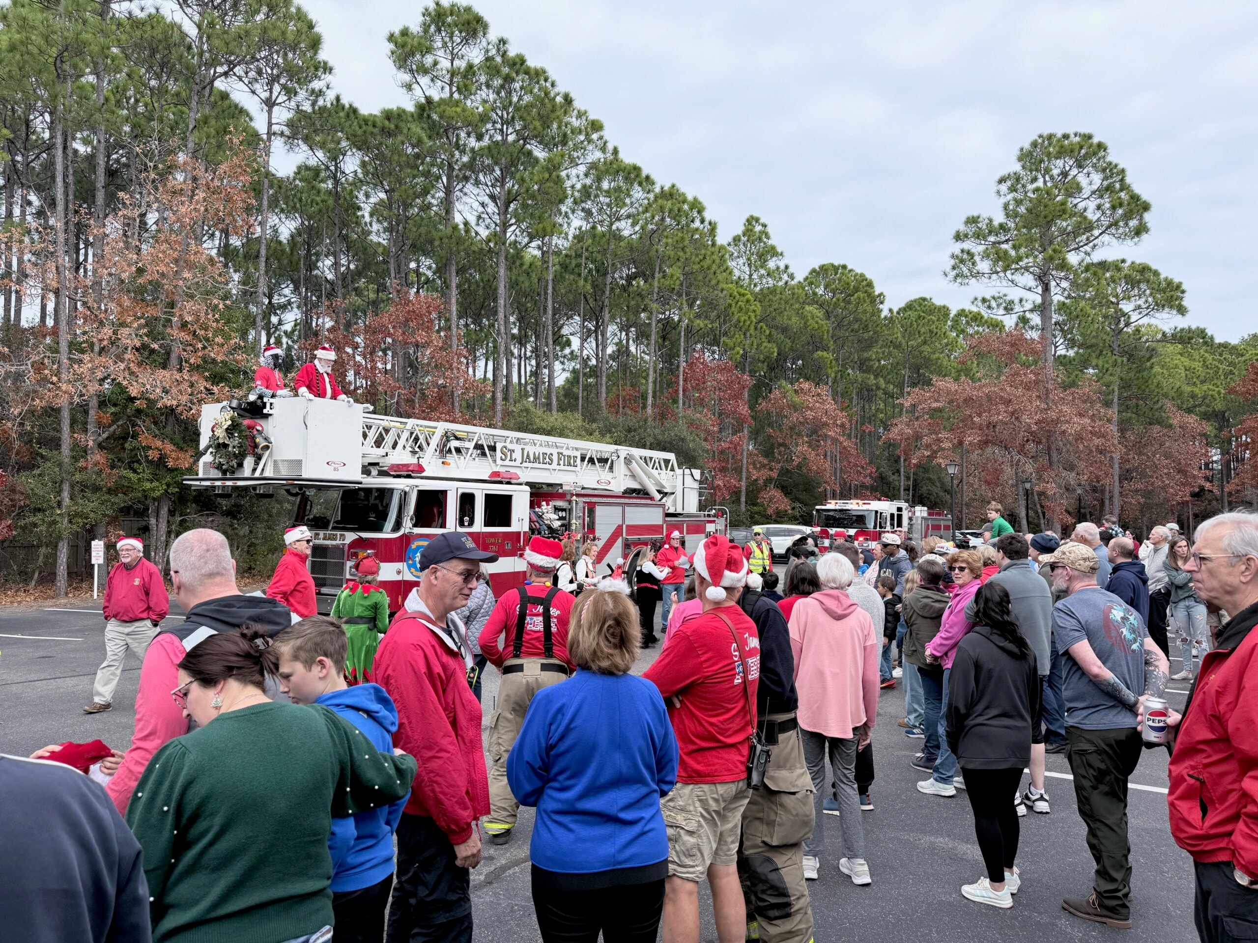 Large crowd of St. James community members gathered in the parking lot as Santa waves from atop the decorated SJFD aerial ladder truck