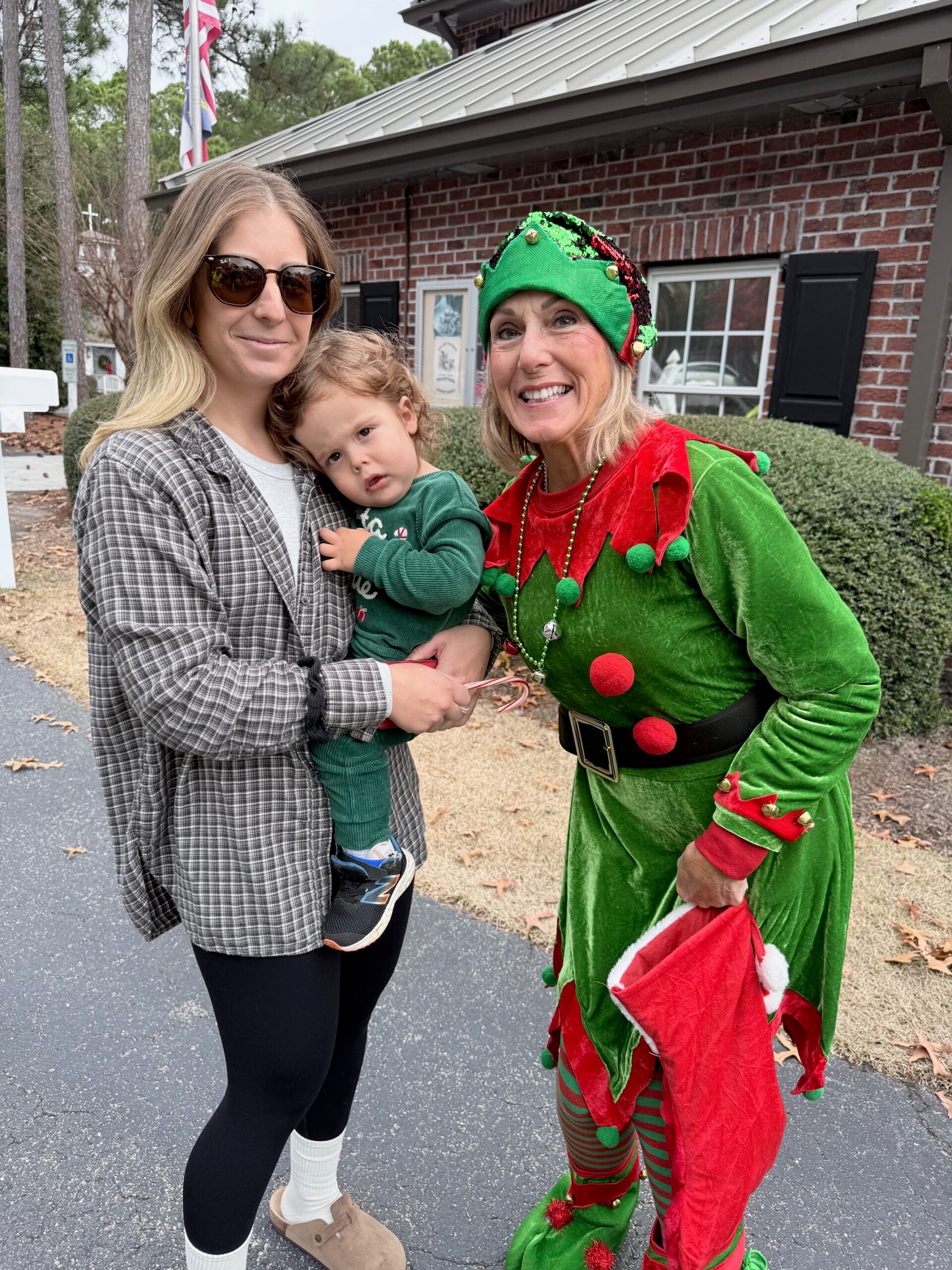 SJFD volunteer dressed as an elf handing a candy cane to a toddler held by his mother outside the St. James Fire station